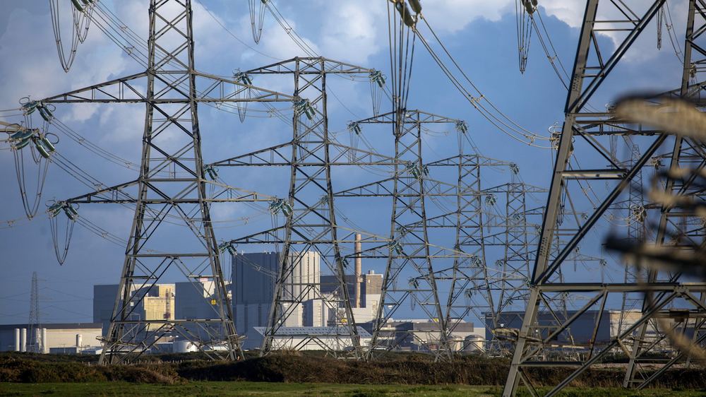 Electricity pylons and power lines in front of the Dungeness Power Station buildings under a cloudy sky.