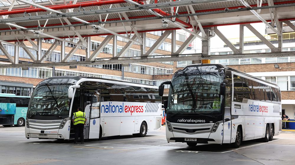 Two National Express coaches parked inside Victoria coach station. A person in a yellow vest stands near one coach's door.