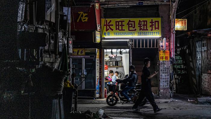 Two workers sit inside a brightly lit workshop at night in the Datang district of Guangzhou, China while a person walks past and another rides a scooter outside in a dimly lit street