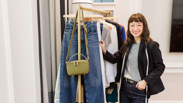 Sera Murphy, smiling, dressed in jeans, grey sweater and black jacket, and with a tape measure draped over her shoulders, stands next to a rail of clothing