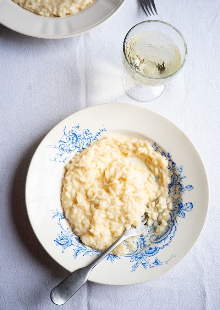 View from above of a plate of pale and creamy risotto, with a fork in the food, alongside a glass of white wine.
