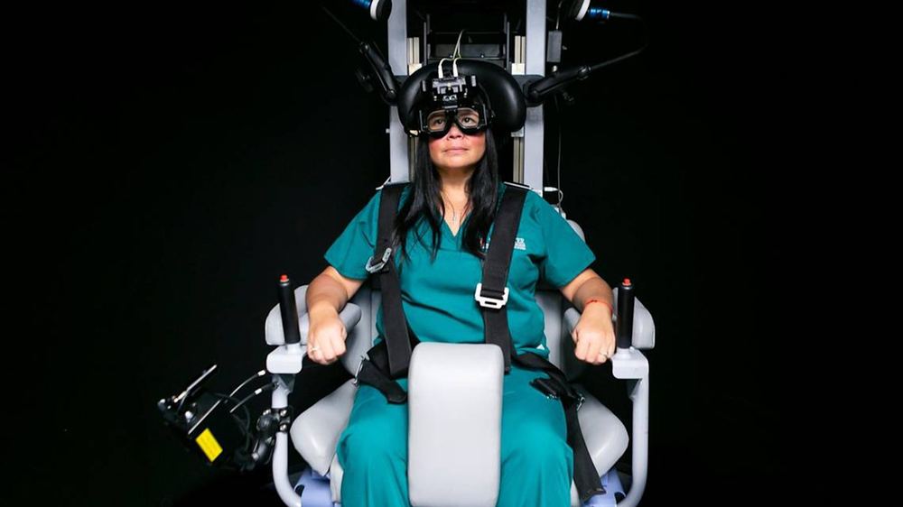 A woman sits in a rotary chair, wearing medical scrubs and diagnostic headgear, used for neurosensory testing.