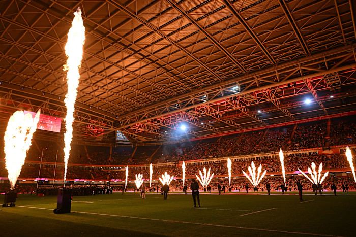 Tall jets of fire shoot upwards from the field as part of a pyrotechnic display before a rugby match at a packed stadium.
