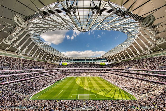 Wide interior view of Tottenham Hotspur Stadium during a football match, with packed spectator stands surrounding the pitch beneath a sweeping roof.