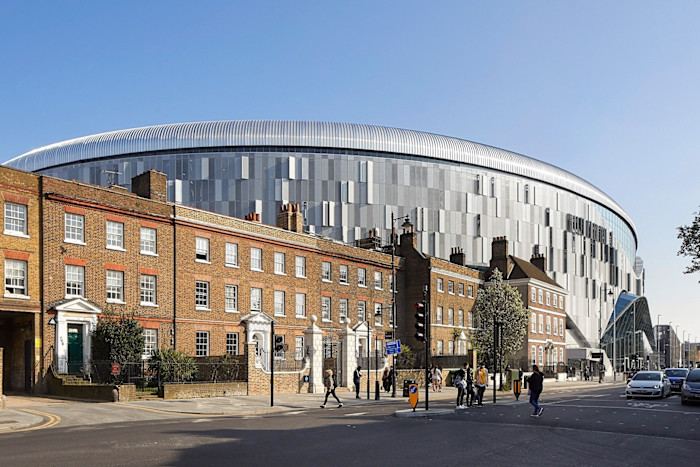 Exterior view of Tottenham Hotspur Stadium in north London, its metallic façade and sweeping roof contrasting with neighbouring historic brick buildings.