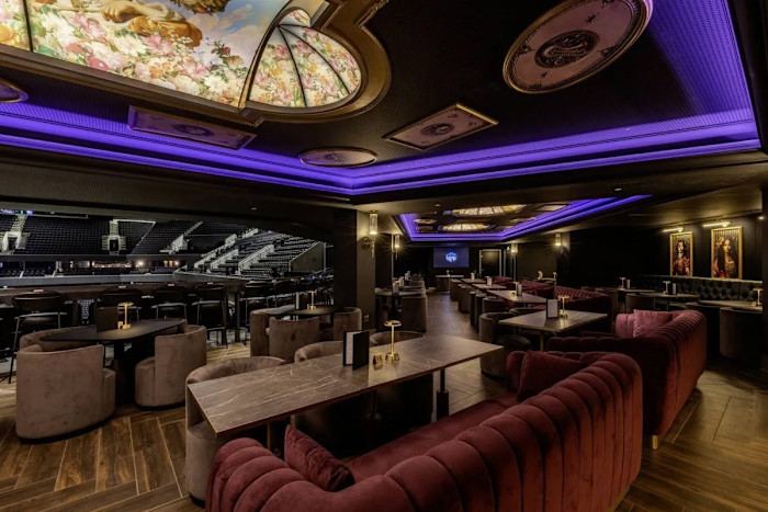 Luxury hospitality lounge overlooking an indoor arena features burgundy velvet sofas, marble tables, purple ceiling lighting and decorative floral ceiling panels.