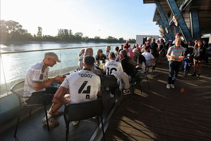 Fans gather on Fulham Pier, some wearing Fulham jerseys, enjoying drinks and the riverside atmosphere before the match.