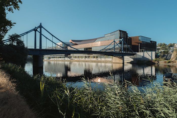 A view of a large modern building with a bronze-coloured facade and a small suspension bridge in the foreground spanning a narrow stretch of water.