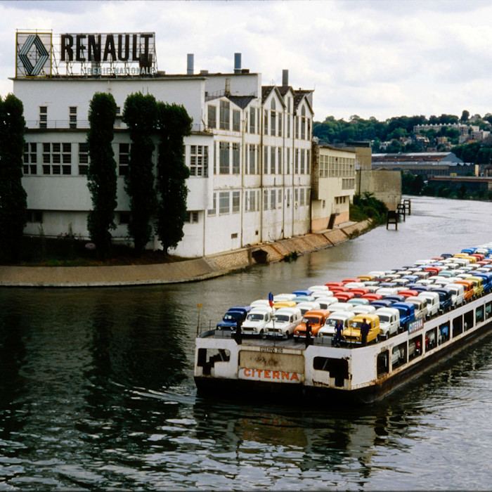 A photograph dated 1980 shows an industrial barge carrying dozens of Renault 4 vans in various colours sailing on the river past a factory with whitewashed walls, chimneys and a giant Renault sign and logo.