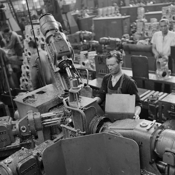 A black-and-white photograph of a male factory worker in a black top and apron operating large-scale machinery.