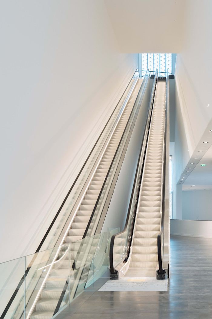 A modern interior of blank gleaming white with an escalator ascending towards a square of light.