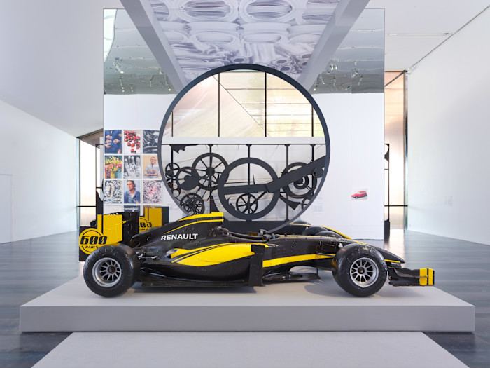 A black-and-yellow Formula 1 racing car on display in an exhibition space, with an image of clockwork and a collage of photographs behind it.