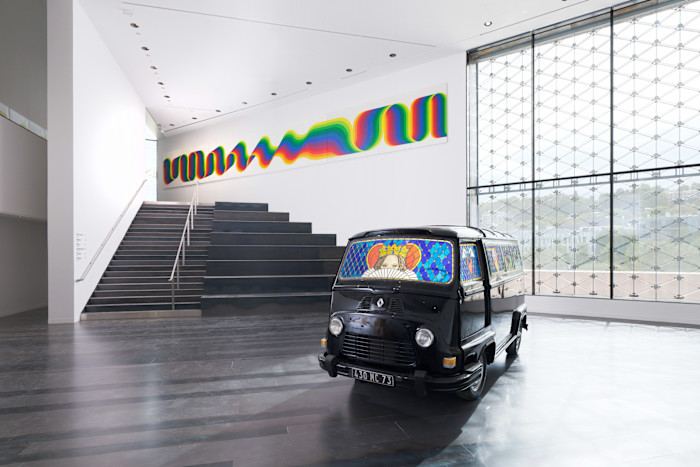 A black 1960s-style Renault van with colourful stained-glass images in its windscreen on display in an exhibition space. There is a large abstract work of art on the far wall in the shape of a wavy rainbow. 