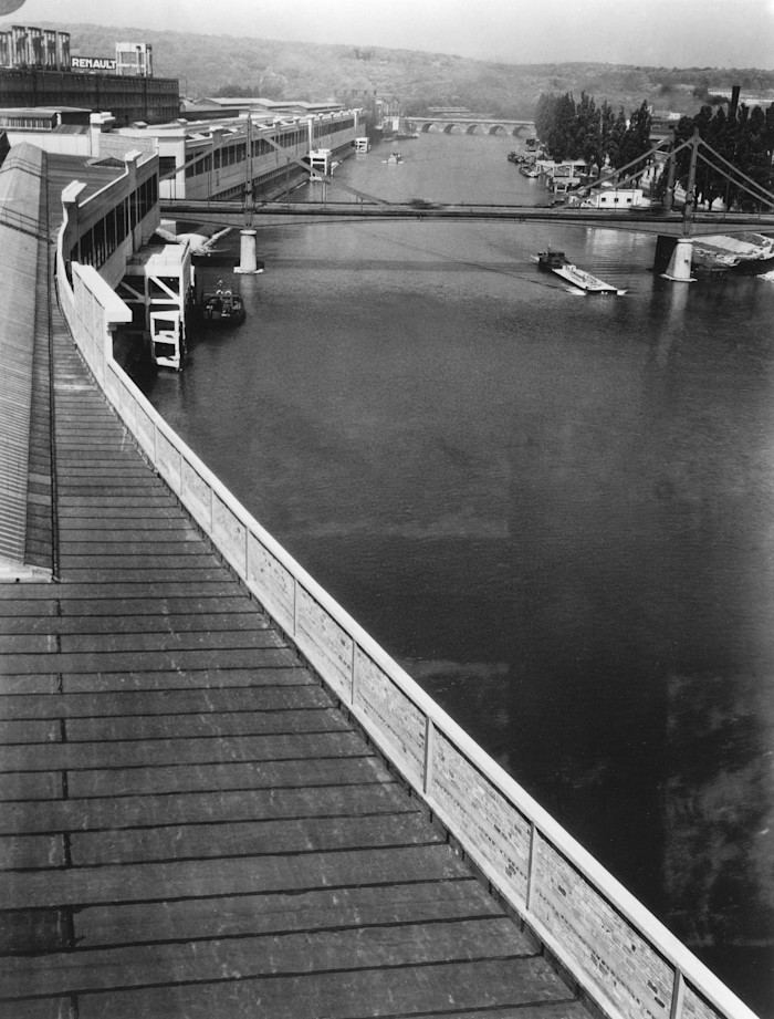A black-and-white photo taken from the roof of a large factory on a curving riverbank, with a view of the Seine, a bridge and distant factory buildings. 