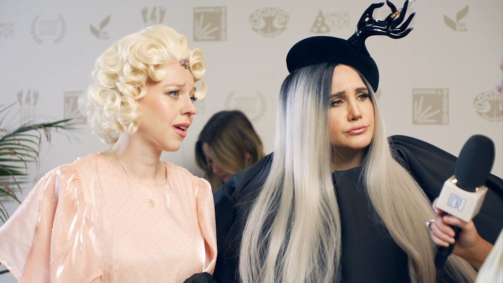 Ellie White and Natasia Demetriou stand together at an event, with a microphone held out towards them.