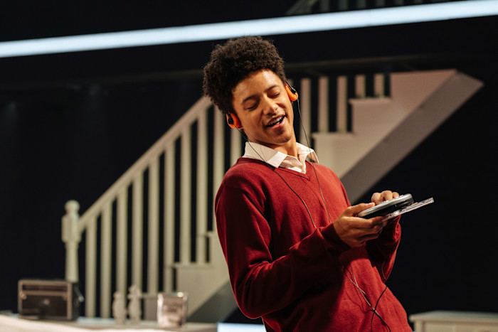Leo Whitfield Harris wears headphones and smiles while holding a portable CD player on stage in ILFORD BOY at Donmar Warehouse.