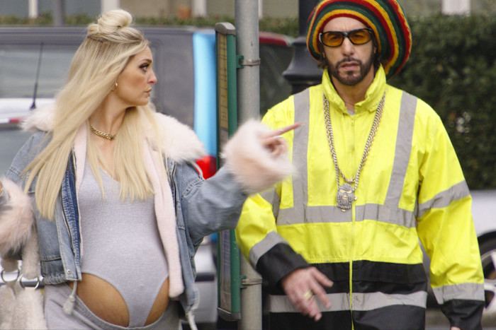 Morgana Robinson as Julie, visibly pregnant, gestures toward Sacha Baron Cohen as Ali G, who wears a neon jacket and colorful hat.