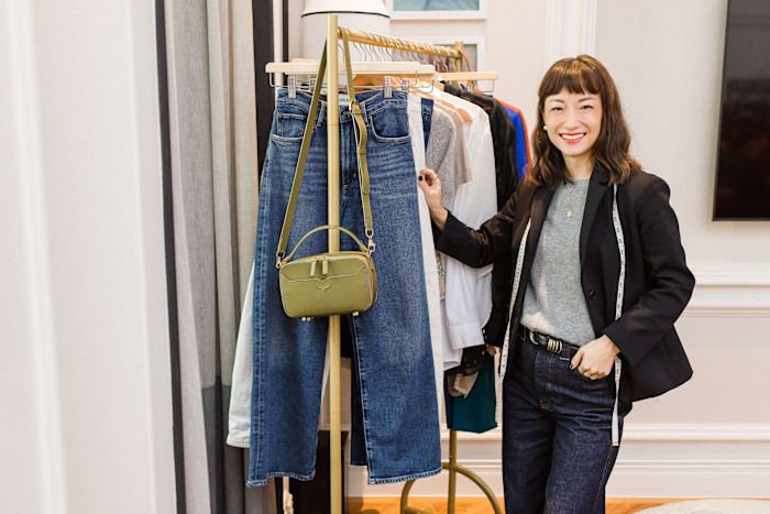 Sera Murphy, smiling, dressed in jeans, grey sweater and black jacket, and with a tape measure draped over her shoulders, stands next to a rail of clothing.
