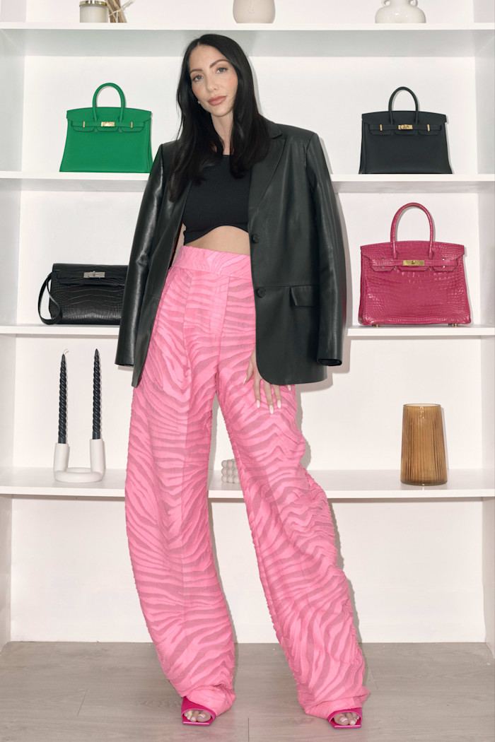 Amber Gordon, wearing pink trousers and black cropped top, with a black jacket over her shoulders, stands in front of shelves on which handbags and candlesticks are displayed.