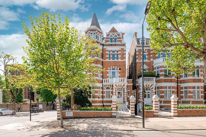 Victorian-style red brick apartment building with white stone detailing, bay windows, and a turret, surrounded by trees on a sunny street.
