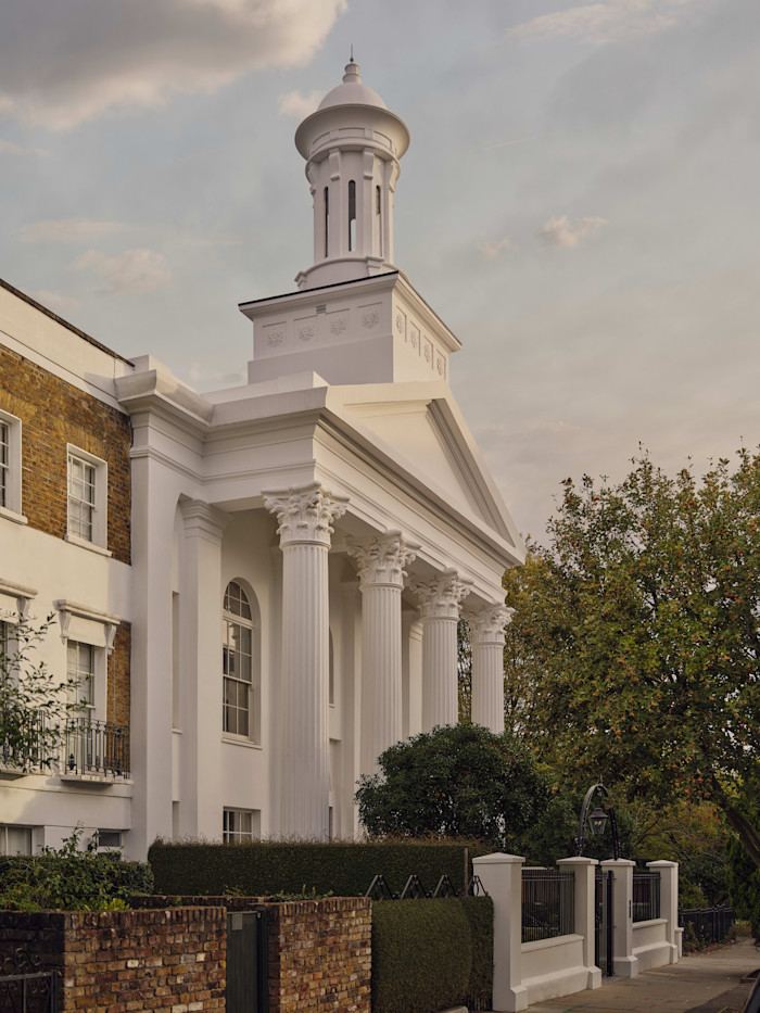 White neoclassical building with tall columns and a distinctive cupola, partially obscured by trees and a brick wall.