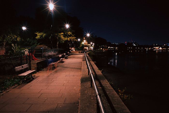 A riverside walkway in London at night, illuminated by street lights, with blurred figures suggesting movement.
