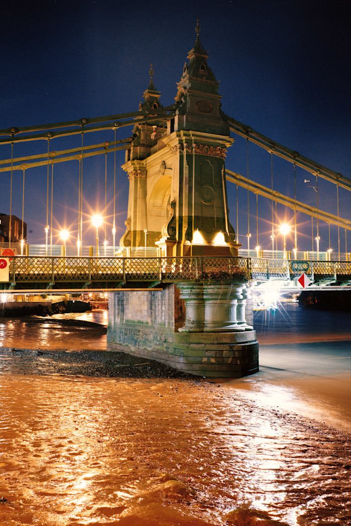 Hammersmith Bridge illuminated at night, with reflections of streetlights on the riverbank below.