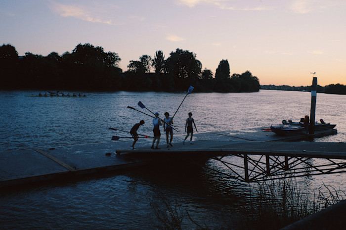 Four people carrying rowing oars stand on a dock at dusk by the river, with a rowing team visible on the water in the background.