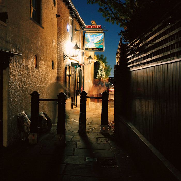 A narrow alley at night leads to a warmly lit pub called The Dove, with a glowing sign and blurred figures walking past.