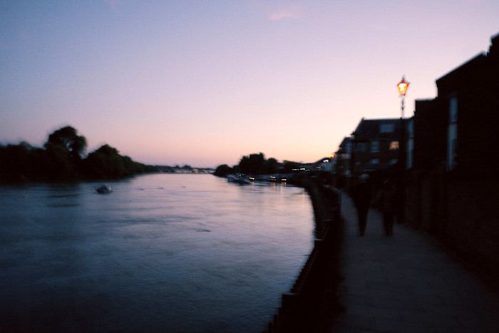 A riverside path in London at dusk with two people walking, street lights glowing and calm water reflecting the evening sky.
