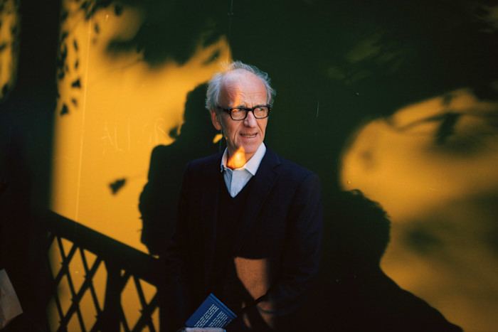 Duncan Minshull in a suit stands against a wall with dramatic yellow and green lighting, holding a book and looking to the side.