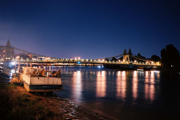 A boat moored on the riverbank at night with Hammersmith Bridge illuminated in the background, reflecting on the water.