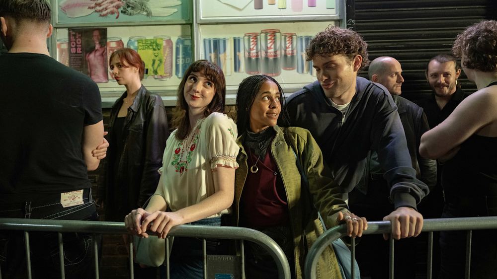 Emily Fairn as Abi, Olive Gray as Sam, and Leo Reich as Ethan stand together in a queue outside at night, leaning on a metal barrier.