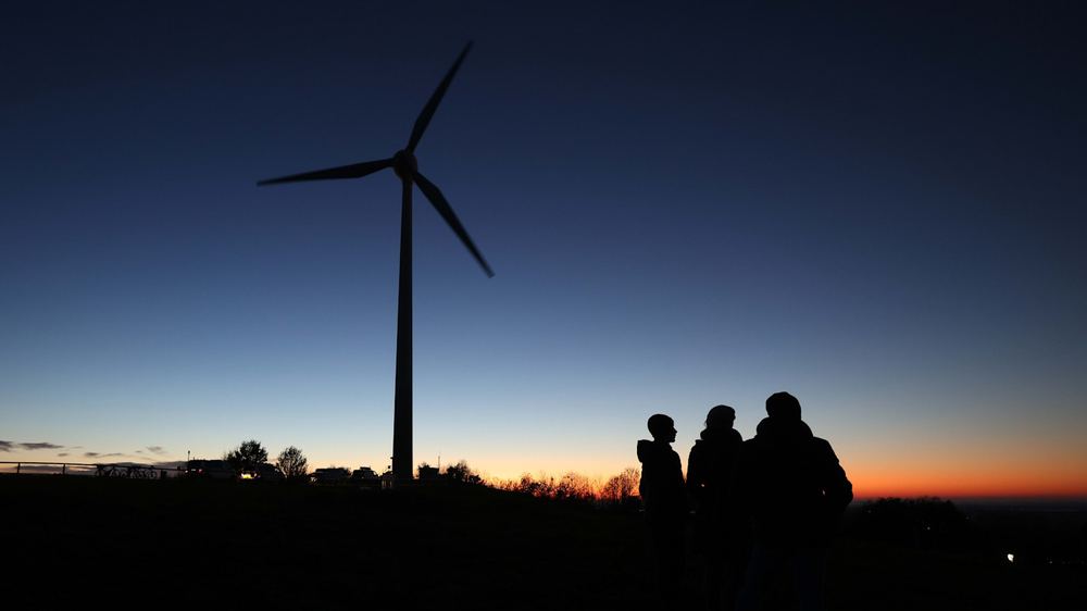 Three people in silhouette stand near a wind turbine at sunset, with a clear sky in the background.