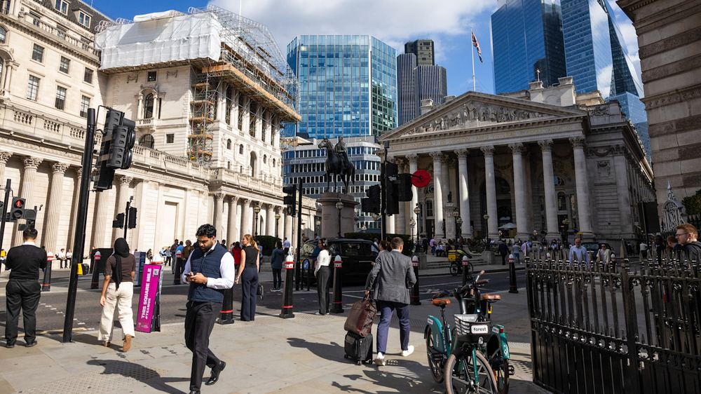 Pedestrians cross a busy intersection outside the Bank of England, with modern skyscrapers towering in the background.