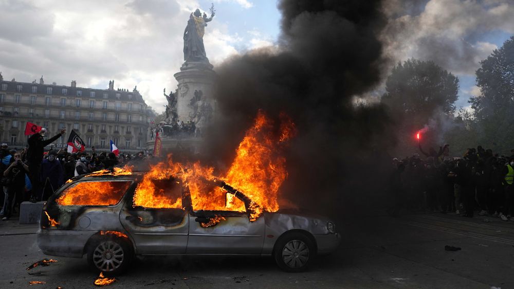 A car engulfed in flames with thick black smoke rises as protesters holding flags and flares gather in République square, Paris.