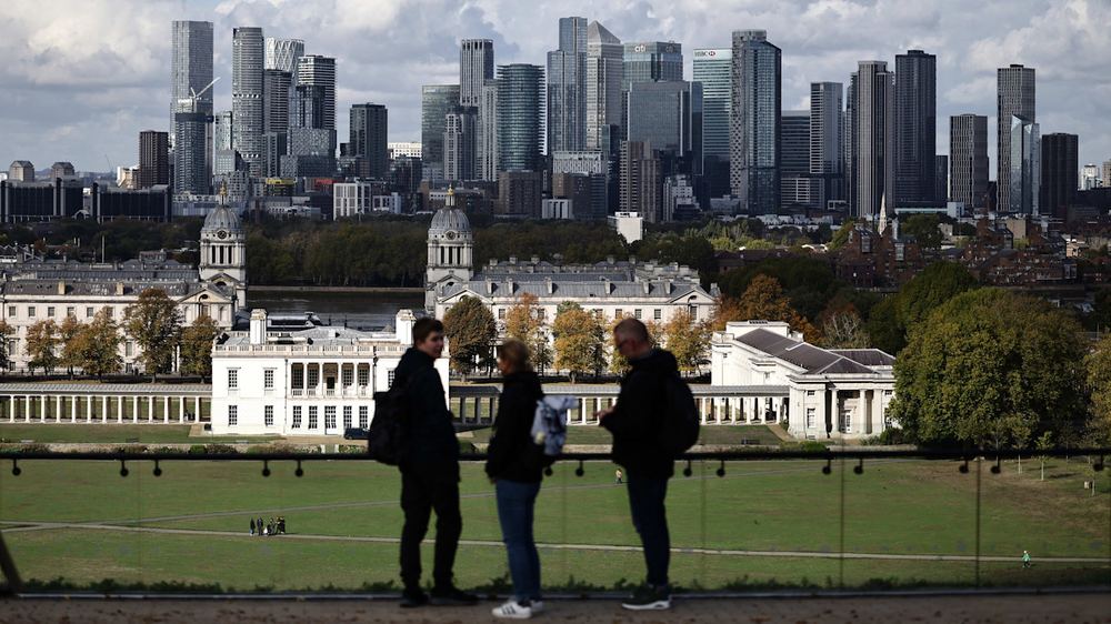 Three people stand in silhouette at a viewpoint in Greenwich Park, with the Old Royal Naval College and Canary Wharf skyscrapers in the background.