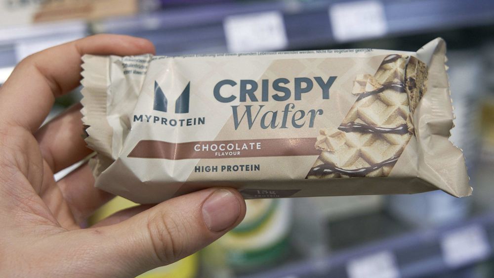 A hand holding a MyProtein Crispy Wafer chocolate high protein snack bar in a supermarket aisle.