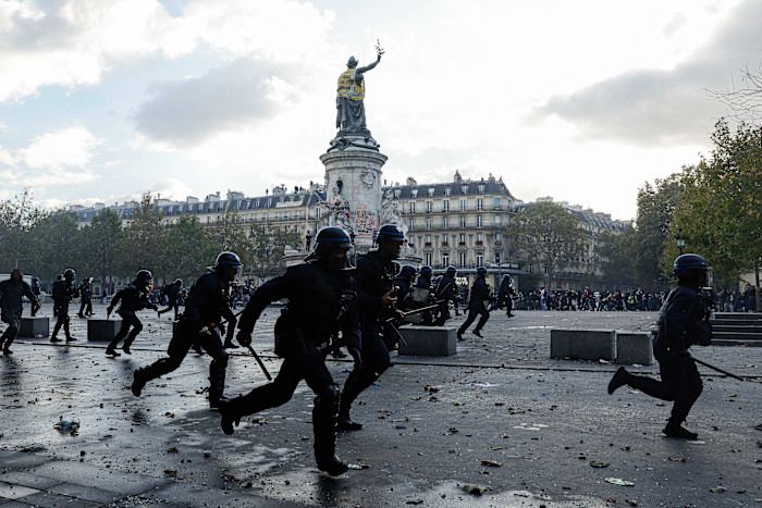 Police officers in riot gear run across Place de la République during a protest, with scattered debris on the ground and the statue in the background.