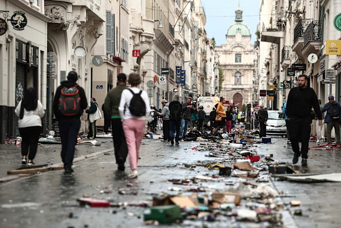 Pedestrians walk along a Marseille street strewn with debris and trash, with damaged storefronts visible after protests.