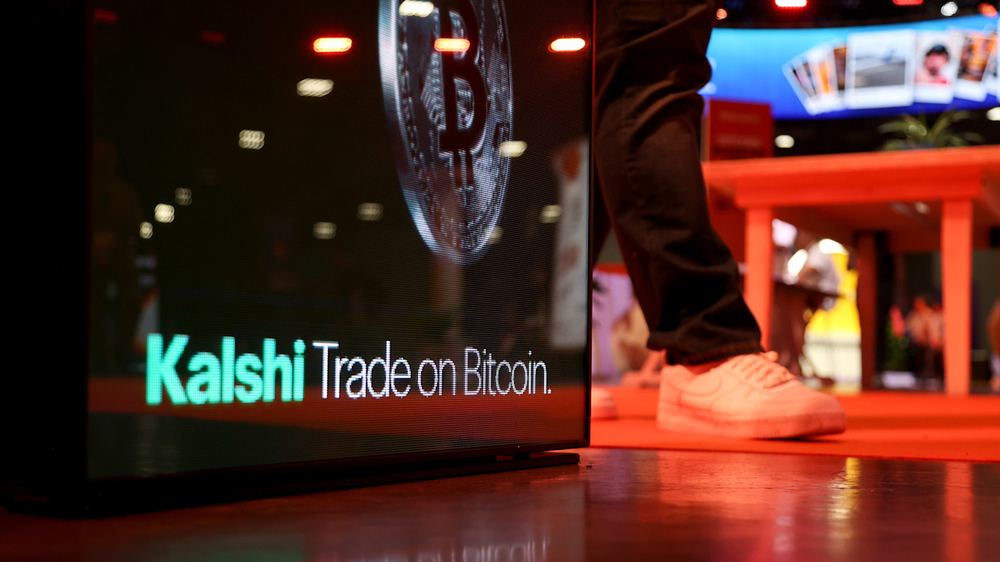 Kalshi sign with the words “Trade on Bitcoin” and a Bitcoin symbol, with a person in white sneakers standing nearby