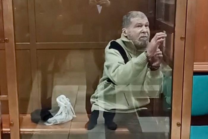 Stephen Hubbard sits in a glass enclosure during a court session, gesturing with his hands.
