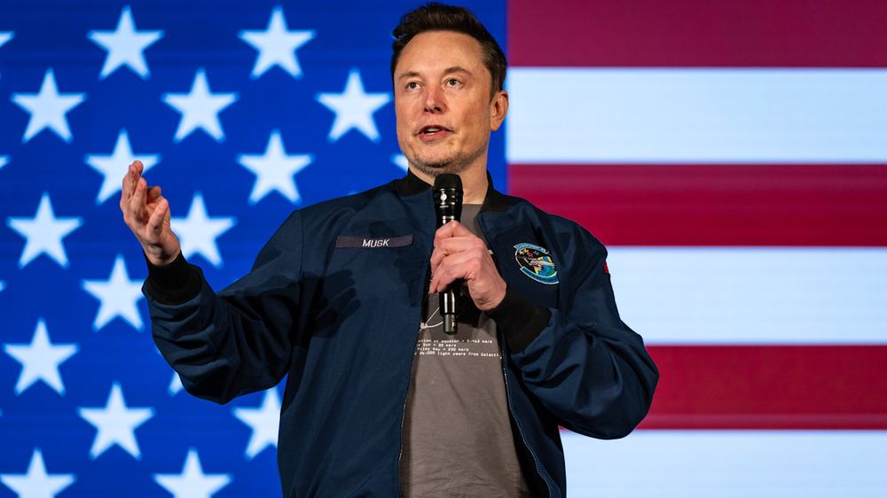 Elon Musk speaks on stage holding a microphone, gesturing with his left hand, in front of a large American flag backdrop.
