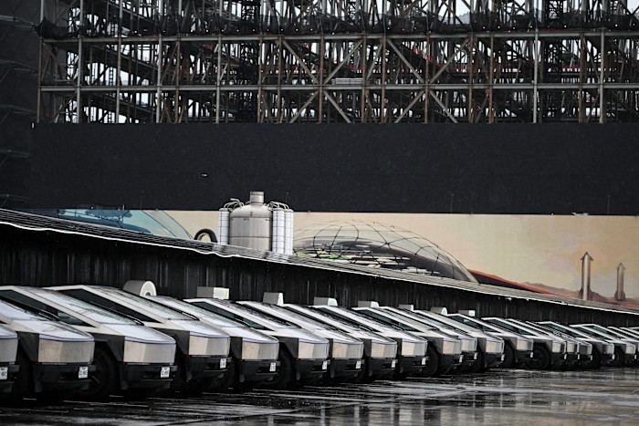 A row of Tesla Cybertrucks is parked in front of a SpaceX facility, with industrial structures and a mural in the background.