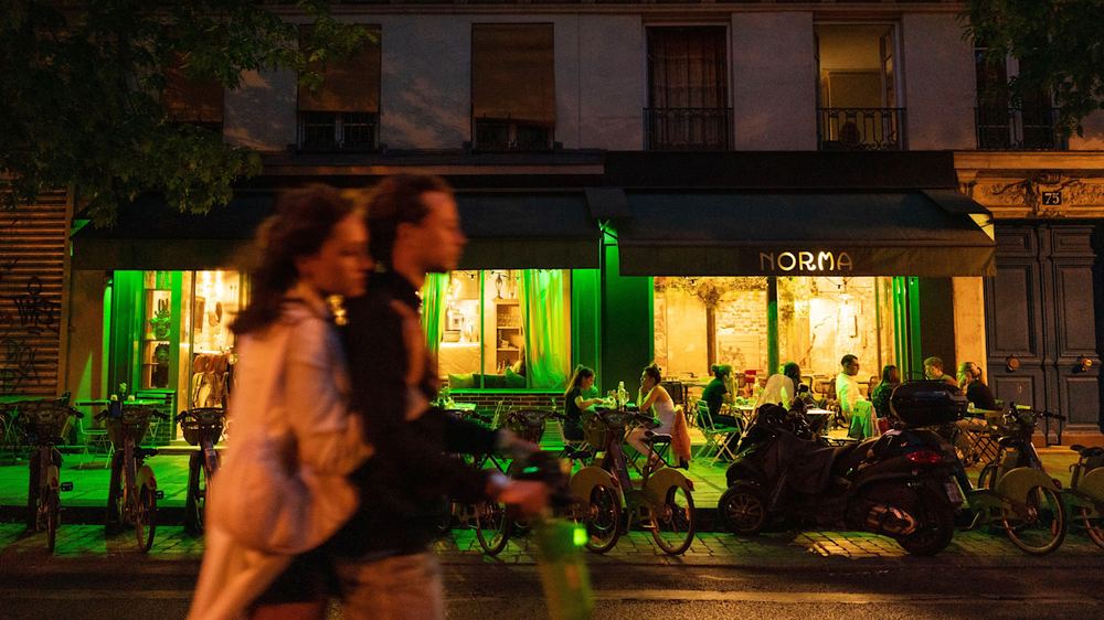 People sit at outdoor tables under green lighting at the restaurant Norma in Paris at night, with blurred pedestrians passing by.