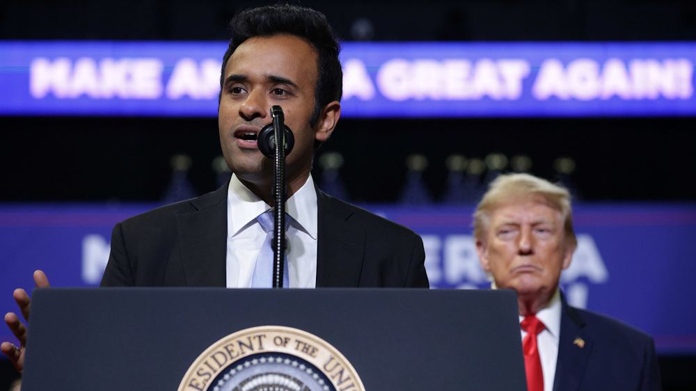 Vivek Ramaswamy speaks at a podium as Donald Trump stands behind him during a rally, with a “Make America Great Again” sign in the background.