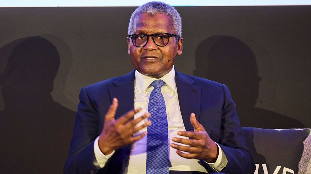 Aliko Dangote speaking on stage, wearing a suit and tie with a headset microphone, gesturing with his hands.