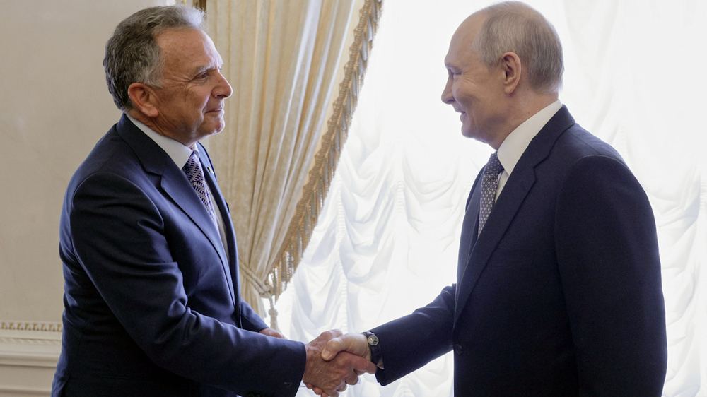 Vladimir Putin and Steve Witkoff shake hands and smile at each other in a room with ornate curtains.
