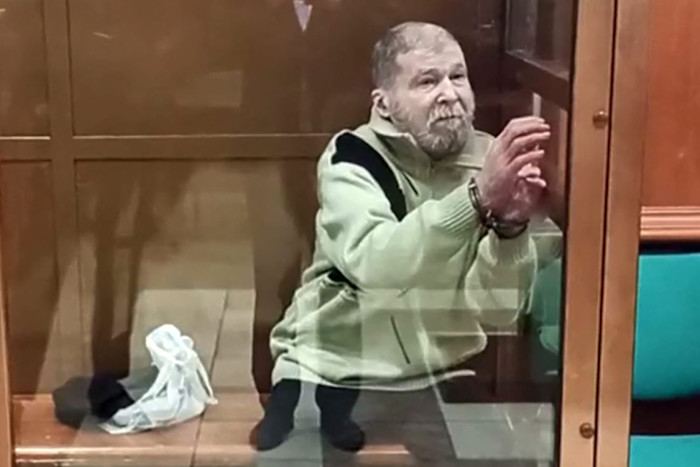 Stephen Hubbard sits behind glass in a courtroom, wearing handcuffs and a light-colored sweater, with belongings on the floor beside him.