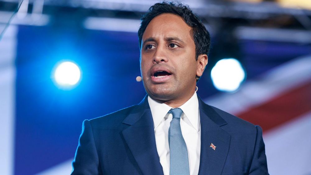 Zia Yusuf speaks at a news conference, wearing a suit and tie with a UK flag pin and a headset microphone.